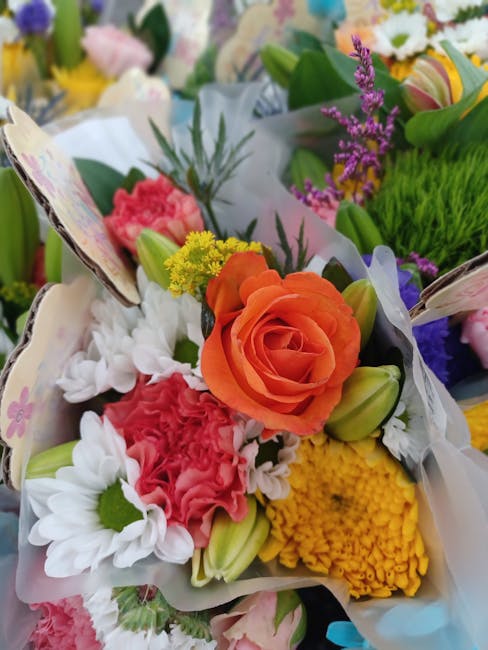 A vibrant floral bouquet featuring a central orange rose surrounded by white daisies, pink carnations, yellow chrysanthemums, purple statice, and green foliage. The flowers are arranged with some buds