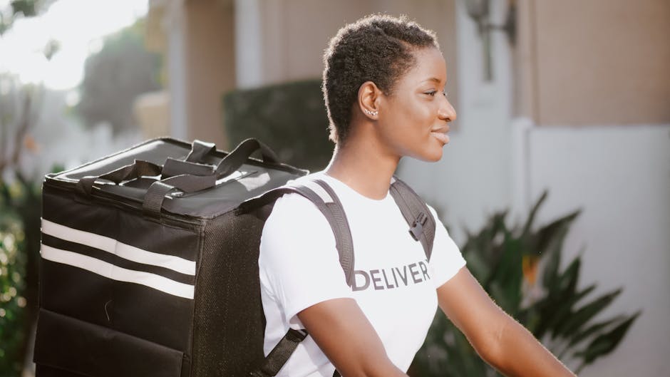 A young woman with short, curly hair and a calm, focused expression is captured outdoors during daylight. She is wearing a white T-shirt with the word 'DELIVERY' printed on it and has a black backpack