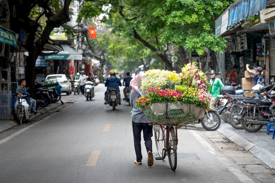 A person pushing a bicycle with a large basket filled with fresh, colorful flowers including pink lilies, white hydrangeas, and yellow roses, on a busy street lined with shops and parked motorcycles. 