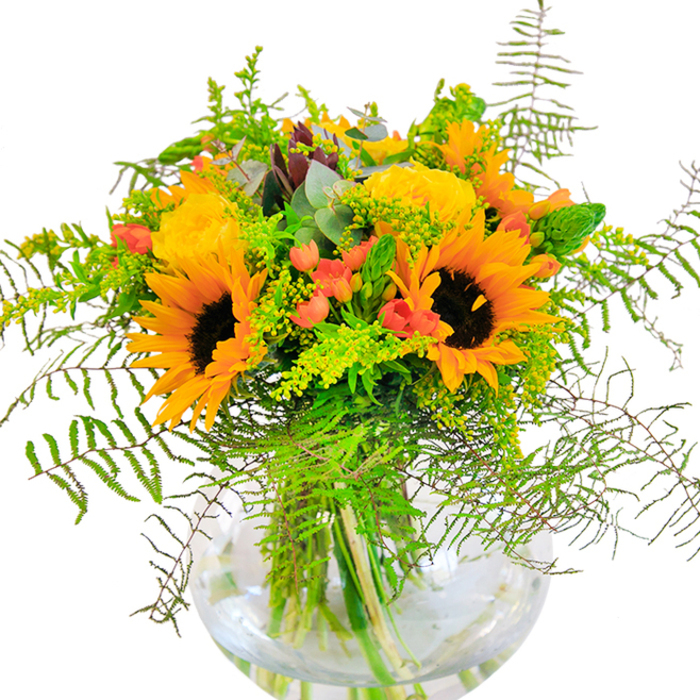 The first impression of this brilliantly bright bouquet is its unapologetic embrace of sunlit exuberance-a signature that mirrors Brompton's graceful, open spaces and blossoming traditions. Shining sunflowers and vibrant yellow roses nestle side by side, their generous, layered petals unfolding to bask in the gentle afternoon light reminiscent of a stroll across Brompton Square. Deep green leaves and feathery, pale green vines spill outward, imitating the lushness of nearby private gardens at their peak. Scattered among the sturdy blooms, vivid scarlet Hypericum berries add a glossy, playful accent to the bouquet's otherwise soft textures, reminiscent of the ripest fruit found at Brompton Road's local markets. The round, clear glass vase cradles the bouquet's architecture, displaying not only the harmonious blend above but also the elegant lines of each stem below, bathed in clear, cool water. Up close, the interplay of textures comes alive: the velvety sunflowers and tightly spiraled roses, the sharp snap of foliage, the smooth coolness of the vase itself. Soft notes of garden scent hint at open meadows and idyllic squares. Designed as a joyful gift marking milestones-birthdays, anniversaries, or times when shared happiness matters-this bouquet brings all the freshness and optimism of a Brompton summer into one radiant arrangement.