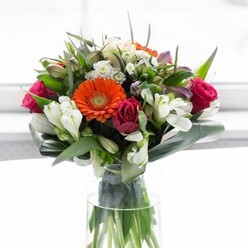 Showcasing a harmonious interplay of colour, structure, and sentiment, this expertly arranged bouquet is a true reflection of Brompton's elegant charm. A halo of creamy, peach-toned gerberas forms a gentle, welcoming embrace around a vibrant cluster of orange petals-each bloom glowing with the cheerfulness of a spring day by Brompton Square. Deep fuchsia roses provide opulent contrast, their tightly packed petals evoking timeless romance, while airy white alstroemeria and petite spray roses pierce through with a touch of brightness and light fragrance. Subtle hints of blush pink and fine, white filler blossoms add depth and fullness, giving the composition an uplifting sense of abundance, as if nature's best were handpicked from local gardens. The robust, glossy aspidistra leaves, carefully curved around the bouquet's base, form a verdant frame that grounds the palette, while slender, pointed green foliage completes the scene with natural grace. The entire arrangement stands tall in a clear glass vase, the intricately woven green stems visible beneath-inviting the eye to appreciate every detail. Sunlight spills delicately across the display, enhancing each hue without overwhelming its gentle beauty. This bouquet was crafted for gatherings and warm gestures, perfect for celebrating a new beginning or simply infusing a Brompton living room with a sense of hope and renewal. Each flower and leaf bears the mark of floral expertise, promising both beauty and emotional resonance.